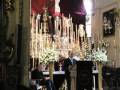 Interior de una iglesia con altar central, flores blancas en el púlpito y personas sentadas en los bancos.