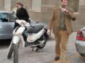 Un hombre camina cerca de una motocicleta aparcada en una calle de ciudad, con una mujer a su lado. La escena muestra un día laboral común en un entorno urbano.