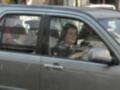 Una mujer con un vestido negro y una corona de flores está sentada en el asiento del pasajero de un coche. La imagen muestra a la mujer sonriendo y mirando hacia el lado del conductor, mientras que el coche parece estar en movimiento.