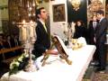 Una imagen de una iglesia con un altar adornado, donde se realizan ofrendas. En el centro hay una mesa cubierta con mantel blanco, sobre la cual se colocan ofrendas y velas. Algunos hombres vestidos formalmente observan el altar, mientras otros están en la parte posterior de la imagen. La decoración interior incluye elementos religiosos y ornamentos dorados, creando un ambiente solemnemente reverente.