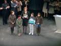 Una escena formal con varias personas, incluyendo adultos y niños. Un niño sostiene un trofeo mientras otros observan con interés. La escena parece ser en una ceremonia o evento formal, posiblemente un concurso o premiación.