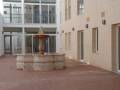 Fotografía de un patio interior con una fuente antigua en el centro, rodeada por edificios modernos con ventanas y puertas de cristal. El suelo es de baldosas rojas, y la atmósfera es tranquila y bien iluminada.