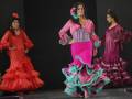 Tres modelos desfilan en trajes de flamenca coloridos y tradicionales, destacando la belleza y elegancia del estilo.