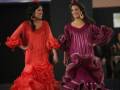 Dos modelos desfilan con vestidos flamencos tradicionales, uno en rojo y otro en morado, en un evento de moda.