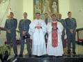 Una imagen de una ceremonia religiosa en un interior con elementos decorativos. En el centro, dos sacerdotes vestidos con hábitos blancos y rojos, uno de ellos con una cruz en la camisa. A su izquierda, un hombre en uniforme militar mira hacia el frente y a la derecha, otro hombre en uniforme militar también mira hacia el frente. En el fondo, se pueden ver cuadros religiosos y una decoración de la pared.