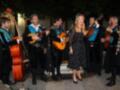 Una banda de música tradicional en un ambiente festivo. Los músicos tocan guitarra, bajo eléctrico y una flauta. La mujer en el centro lleva un vestido negro con lunares blancos y tiene una corona de flores. La escena parece ser al aire libre, posiblemente en una fiesta o evento cultural.