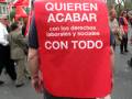 Un hombre con una chaqueta roja y un mensaje que dice "QUIEREN ACABAR con los derechos laborales y sociales CON TODO" en una manifestación.