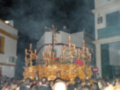 Una procesión religiosa nocturna con una imagen de Cristo crucificado en un manto dorado, rodeada por velas encendidas y una multitud de fieles observando.