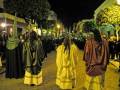 Una procesión nocturna con personajes vestidos de penitentes, en una calle adoquinada. La imagen muestra a varias personas con capas de diferentes colores, incluyendo verde, marrón y amarillo. Algunos portan máscaras que cubren la cara, mientras otros llevan capuchas largas. La escena está iluminada por faroles y luces de la calle, creando un ambiente festivo. En el fondo se observan edificios con árboles decorativos.