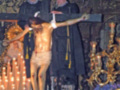 Imagen de un Cristo crucificado en una iglesia, rodeado por dos hombres vestidos con trajes ceremoniales. La imagen está iluminada por velas y adornos dorados en el fondo, creando un ambiente de solemnidad.