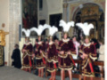 Imagen de una procesión religiosa en un templo, con personas vestidas con trajes tradicionales de color rojo y dorado, adornadas con plumas blancas. La escena se desarrolla en un ambiente histórico y religioso, destacando la belleza de los trajes y la solemnidad del evento.