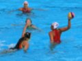 Jugadores de waterpolo en acción, con una jugadora en rojo y otra en verde intentando bloquear el balón.