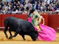 Un torero con capa verde y sombrero negro enfrenta a un toro negro en una plaza de toros. La escena está rodeada por espectadores y un muro rojo en el fondo.