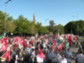 Una multitud de personas con pancartas y banderas se concentra frente a un edificio histórico, bajo un cielo azul claro. La imagen muestra una manifestación o protesta en un lugar público, con árboles y edificios al fondo.