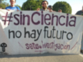 Un grupo de personas sostiene un cartel blanco con letras negras y moradas que dice "#sinCiencia no hay futuro" en español. El cartel también contiene la frase "setas@investigación" en letras más pequeñas y rojas. La imagen muestra a varias personas con pancartas, lo que sugiere una manifestación o protesta.