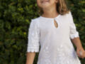 Una niña con cabello largo y ondulado lleva un vestido blanco bordado, posando frente a una pared de hiedra.