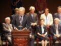 Un grupo de personas en trajes formales se encuentran en un escenario, con uno de ellos al frente de un podium. La imagen parece ser de una ceremonia o evento importante, posiblemente un premio o reconocimiento.
