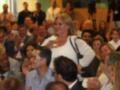 Una mujer con una camisa blanca y un bolso negro está entre una multitud de personas en un evento. La imagen muestra a varias personas con diferentes expresiones y atuendos, lo que sugiere una reunión o conferencia.