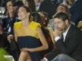 Una mujer con un vestido amarillo y azul está sentada junto a un hombre en una ceremonia, ambos sonríen. La imagen muestra a varias personas en trajes formales sentadas en filas, con copas de champán y luces de fondo.