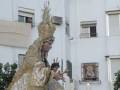 Imagen de una procesión religiosa con una imagen de la Virgen María vestida en traje dorado, sosteniendo un niño y rodeada de velas. La escena se desarrolla en un entorno urbano con edificios blancos y ventanas.