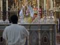 Una persona vestida con un hábito blanco se encuentra frente a una mesa altar decorada con velas y objetos religiosos, en un ambiente que parece ser una iglesia o templo.