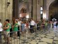Una multitud de personas espera en fila para entrar a una catedral, con arcos y columnas de piedra. La imagen muestra la entrada principal de la catedral, con gente organizada en filas y una valla metálica para separar la fila de los visitantes.