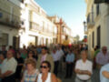 Una multitud de personas camina por una calle, entre edificios blancos y rojos. La gente parece estar en un evento o celebración, con algunos hombres vestidos formalmente y mujeres con sombrillas. La imagen muestra un ambiente festivo en una ciudad típica.