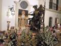 Una procesión religiosa en un templo, con una estatua de oro y plata que parece representar a un santo o personaje importante. La estatua está sobre una plataforma dorada con flores blancas y un fondo de madera pintada. La imagen también incluye a personas en trajes tradicionales, posiblemente fieles o participantes de la procesión. En el fondo, se puede ver una estructura con un arco y un escudo, probablemente simbolizando el emblema de la ciudad o región.