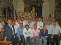 Grupo de hombres en una iglesia, posando para la foto. Algunos llevan camisas y otros corbatas. La imagen muestra detalles de la decoración interior de la iglesia, incluyendo un altar y vitrales.