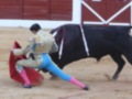 Un torero en traje tradicional lucha con un toro negro en una plaza de toros. El fondo muestra una pared roja y blanca con un diseño geométrico.