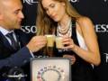 Una modelo y un presentador toman café con una máquina de Nespresso en la mano. El fondo es negro con el logo de Nespresso visible.