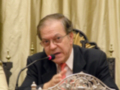 Un hombre con cabello gris y gafas, vestido de negro y corbata roja, está sentado en una mesa con un micrófono frente a él. La imagen parece ser de una conferencia o reunión formal, con cortinas en el fondo y un escritorio ornamental.