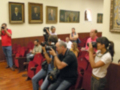 Una persona en rojo toma fotos de un hombre con cámara, mientras otros asistentes observan. El ambiente es formal, posiblemente una sala de conferencias o un salón público.