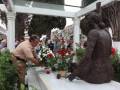 Una estatua de bronce de un hombre sentado en una tumba, rodeada por flores y velas. Un grupo de personas se reúne alrededor, algunas con sombrillas y otros adornos. El entorno es un cementerio con estructuras de madera y vegetación en el fondo.