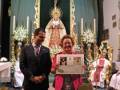Una imagen de una iglesia con un altar en el fondo, donde se puede ver la Virgen María y un crucificado. En primer plano, dos personas están sosteniendo una bandera con el símbolo de la Unión Europea. La persona izquierda lleva un traje negro y corbata, mientras que la derecha tiene un vestido largo con detalles coloridos. La iglesia está decorada con velas y flores, creando un ambiente festivo y religioso.