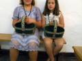 Dos niñas con vestidos blancos y coronas posan con cestas de mimbre en una escena festiva.