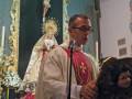 Un sacerdote vestido con un hábito de color claro sostiene una imagen religiosa mientras se encuentra en una iglesia. La imagen muestra una estatua de la Virgen María y un crucifijo en el fondo.