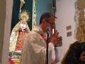 Un sacerdote en vestimenta litúrgica orando frente a una imagen venerada, rodeado de ornamentos religiosos y decoraciones.