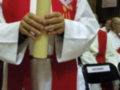 Un sacerdote sostiene una vela amarilla en un entorno religioso, con vestimenta sacerdotal y una saya roja.