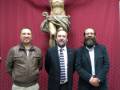 Tres personas posan frente a una estatua de Cristo en la cruz, en un ambiente formal con un fondo de madera.
