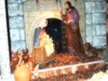 Escena navideña con figuras de madera en un entorno de piedra, incluyendo una escena de la natividad con un bebé Jesús en el centro. A los lados hay figuras humanas y animales, rodeadas de adornos navideños como naranjas y frutas. La imagen está iluminada con luces de navidad, creando un ambiente festivo y tradicional.