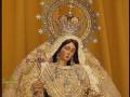 Una estatua de una Virgen María vestida con una elegante vestimenta dorada y perlada, posiblemente en un entorno religioso o festivo. La Virgen lleva una corona y un velo, con joyas y símbolos religiosos en su vestimenta. La escena es simbólica y probablemente representa a una figura de devoción o veneración en un contexto cultural o religioso.