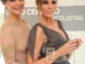 Dos mujeres sonrientes posan juntas en un evento formal, una con vestido beige y la otra con vestido gris. La imagen incluye un fondo con letras en español que parece ser parte de una publicidad o anuncio.