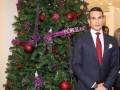 Un hombre en traje negro posa frente a un árbol de Navidad adornado con bolas rojas y purpuras. La palabra "PORCELANO" se puede leer en la cinta de la decoración.