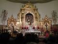 Interior de una iglesia con altar dorado, velas y flores rojas. Personas en la oración.