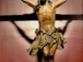 Escultura en madera de Cristo crucificado, con una expresión dolorosa. La cruz está claramente visible detrás de él. El fondo es oscuro, enfocando la figura del Cristo en el centro.