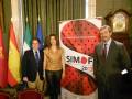 Tres personas posan frente a una bandera española, un cartel de SIMOF 2013 y banderas europeas.