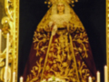 Imagen de una Virgen María vestida con un manto negro y dorado, rodeada por flores blancas y velas encendidas.