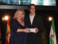 Persona entregando un premio a otra en un evento formal con banderas de España y Andalucía en el fondo.