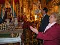 Una mujer con una chaqueta roja y un suéter rosa hablando en un altar dorado. Detrás de ella, hay una figura vestida con velos y un hombre en traje. La imagen muestra detalles de la decoración religiosa, incluyendo velas altas y un altar con flores rojas.